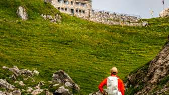 Wanderer mit rotem Rucksack blickt auf die Pfälzerhütte inmitten grüner Berglandschaft