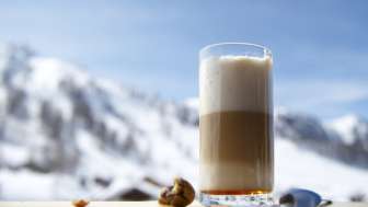 Glas Latte Macchiato auf einer Holzplatte vor der Kulisse verschneiter Berge