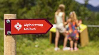 Wegweiser des Alpherzwegs Malbun mit Kindern vor einer der Stationen im Hintergrund