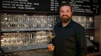 Sympathischer Barkeeper der WY Weinbar mit Weinglas vor stilvollem Regal voller Gläser und Weine.