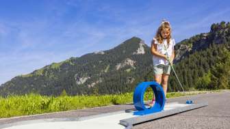 Mädchen konzentriert sich beim Minigolfspielen auf eine blaue Looping-Bahn in alpiner Kulisse