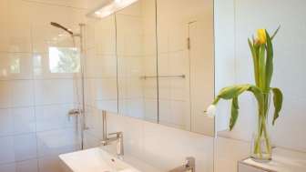 Modernes, hell gefliestes Badezimmer im Gästehaus beim Camping Mittagsspitze mit Doppelwaschbecken, Spiegel und frischen Blumen.