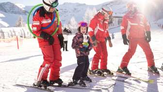 Drei Skilehrer in roten Anzügen und ein Kind auf Skiern im Schnee, im Hintergrund verschneite Berge und Sonnenschein
