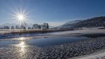 Sonnenaufgang am Egelsee in Mauren im Winter mit glitzernder Eisfläche, Schnee und sonniger Bergkulisse.