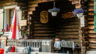 Eingangsbereich der Hütte: Rustikaler Eingangsbereich der Gafadurahütte mit Holzfassade, bunten Blumen und Hinweistafel