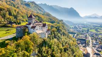 Luftaufnahme von Schloss Vaduz auf einem Hügel über der Stadt, mit Alpenpanorama und Blick ins Rheintal bei klarer Sicht.
