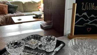 Kristallschalen auf einer Küchentheke vor einem gemütlichen Wohnbereich und eine Tafel mit der Aufschrift «Herzlich Willkommen»
