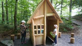 Zwei Kinder erkunden eine kleine Holzhütte im Wald, die Teil des Erlebniswegs „Wurz und Zirp auf Zeitreise“ in Liechtenstein ist – ein Naturabenteuer zum Anfassen.